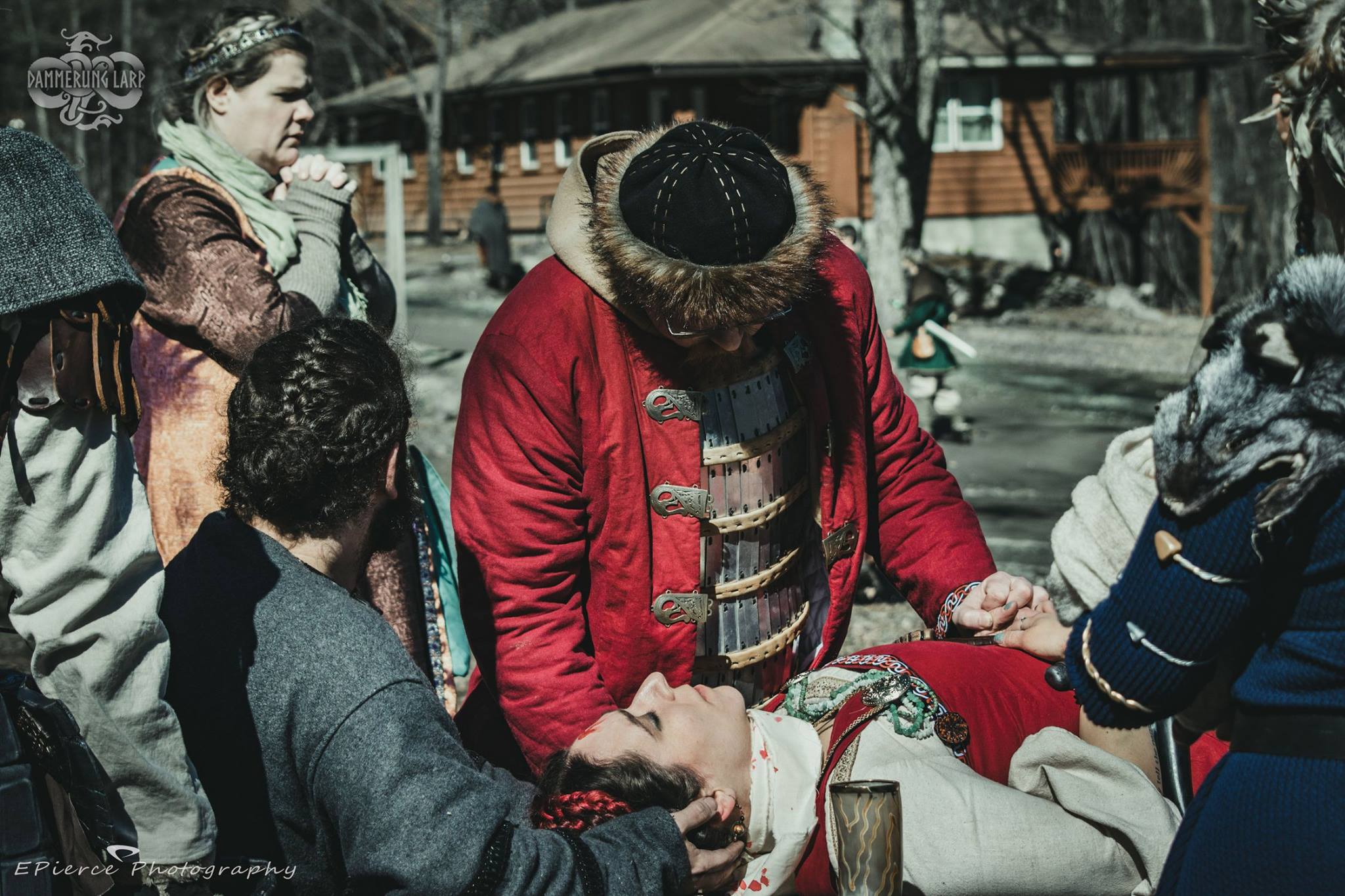 A man in viking clothing holds the hand of an unconscious, bloodied woman on a table as their religious adviser looks on.