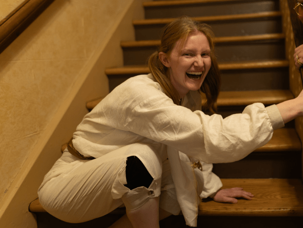 A woman in white crouches on a staircase with a manic laugh on her face.